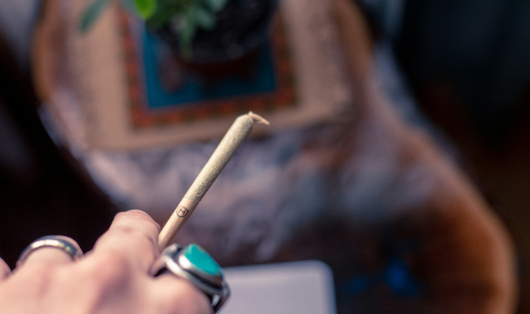 Hand holding a lit cannabis joint with smoke in background